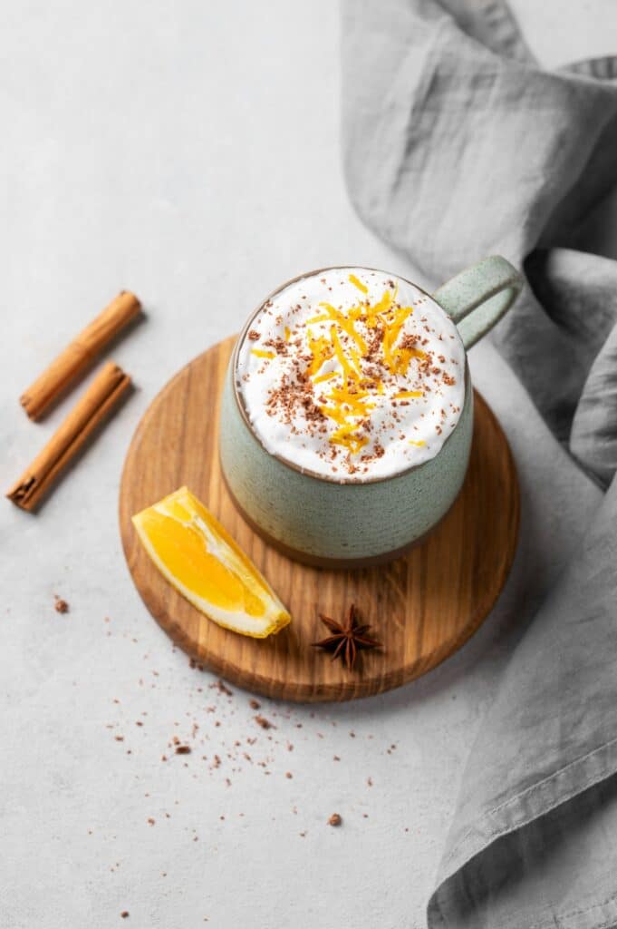 Mug of hot chocolate with whipped cream and orange zest on a wooden board on a light background